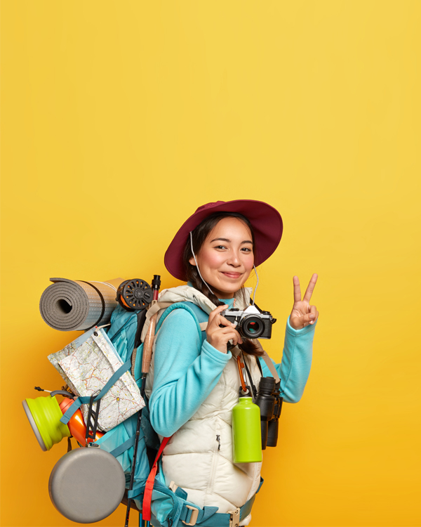 active-female-traveler-makes-peace-gesture-holds-retro-camera-taking-pics-carries-big-rucksack-with-destination-map-karemat-other-tourist-things